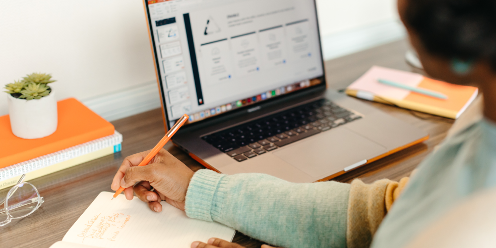 A person writes down notes while looking at a laptop screen.