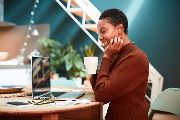 a woman holds a mug and smiles while working on a laptop.