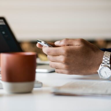 Close up of someone's hands holding a mobile device above a desk with a laptop in the background.