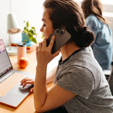 A woman talks a cell phone while scrolling on a laptop.