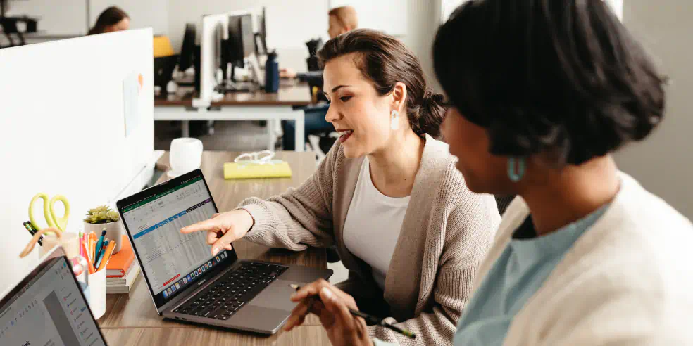 Two coworkers seated side by side discussing something on one of the laptops in front of them.