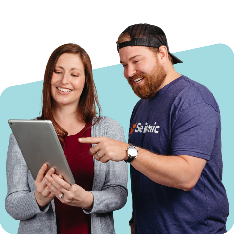 Two smiling people standing side by side. One of them is holding a tablet, the other is pointing to it.