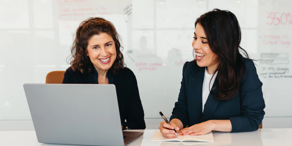 Two smiling women looking at laptop. One of the them is writing notes in paper notebook.