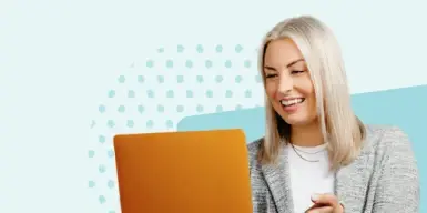 A smiling woman, looking at a laptop, engaged in an online meeting.